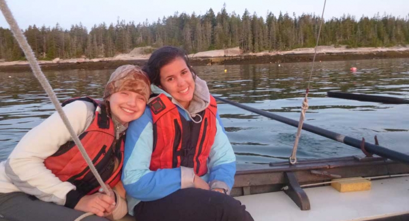 Two people wearing life jackets sit closely and smile for the photo. They are on a boat on calm water. 
